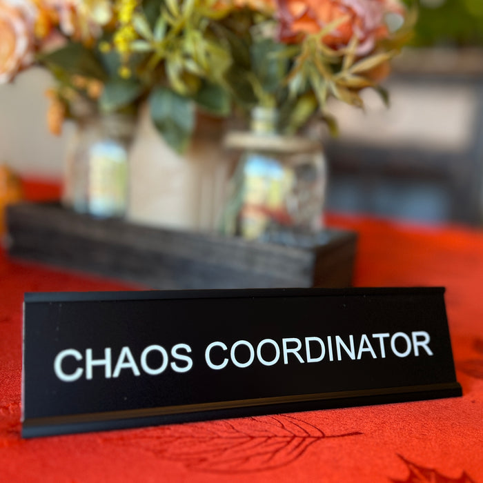 Funny desk sign reading 'CHAOS COORDINATOR' in front of a blurred floral arrangement.
