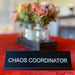 Table sign reading 'Chaos Coordinator' on a red tablecloth with a blurred background of flowers and decor.