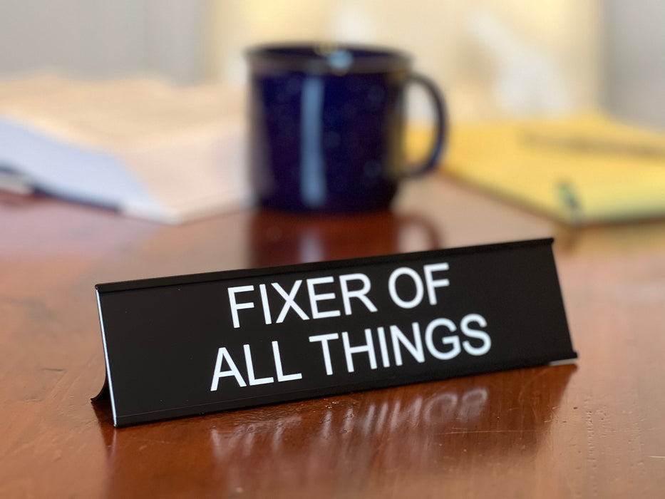 Funny office decor fixer of all things desk sign sitting in front of a coffee mug.
