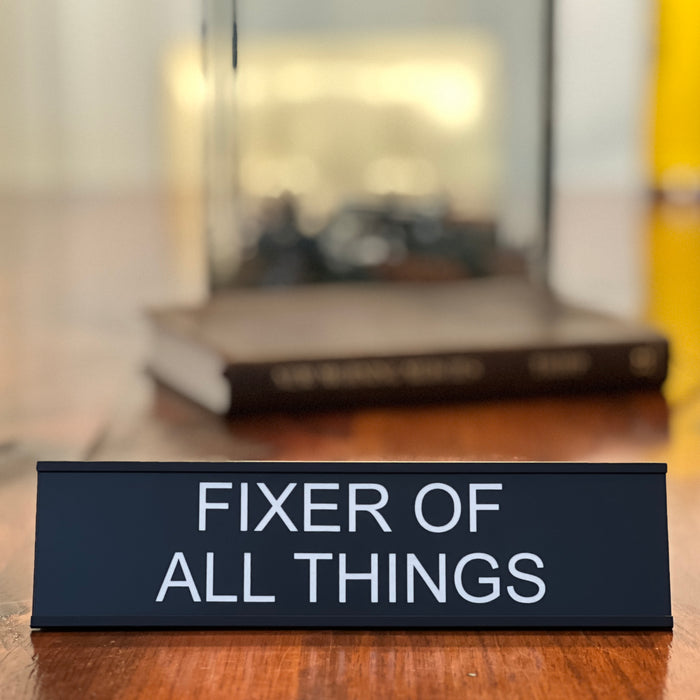 Funny office decor fixer of all things desk sign sitting in front of a book.
