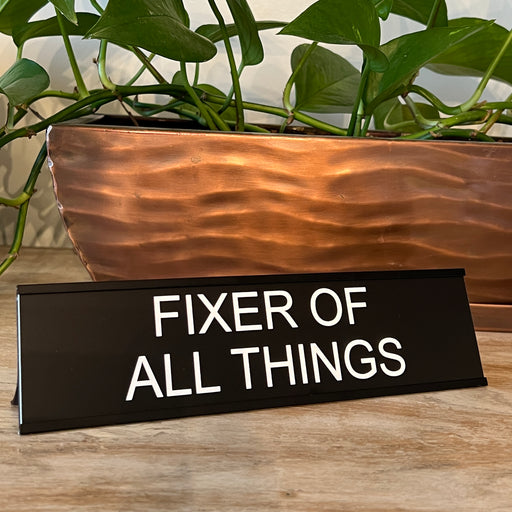 Fixer of all things funny office desk plate in front of a plant.
